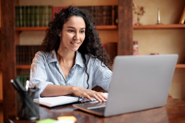 Doing Project. Portrait of curly young woman using laptop computer and typing on keyboard, writer sitting at table. Female manager working remotely or studying online at home, telecommuting